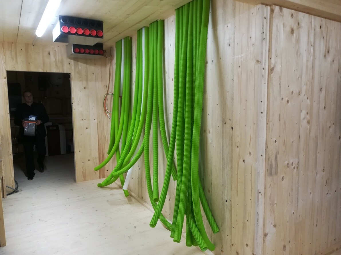 Green flexible hoses hanging from the ceiling along a wooden wall; a man stands in the doorway holding a box.
