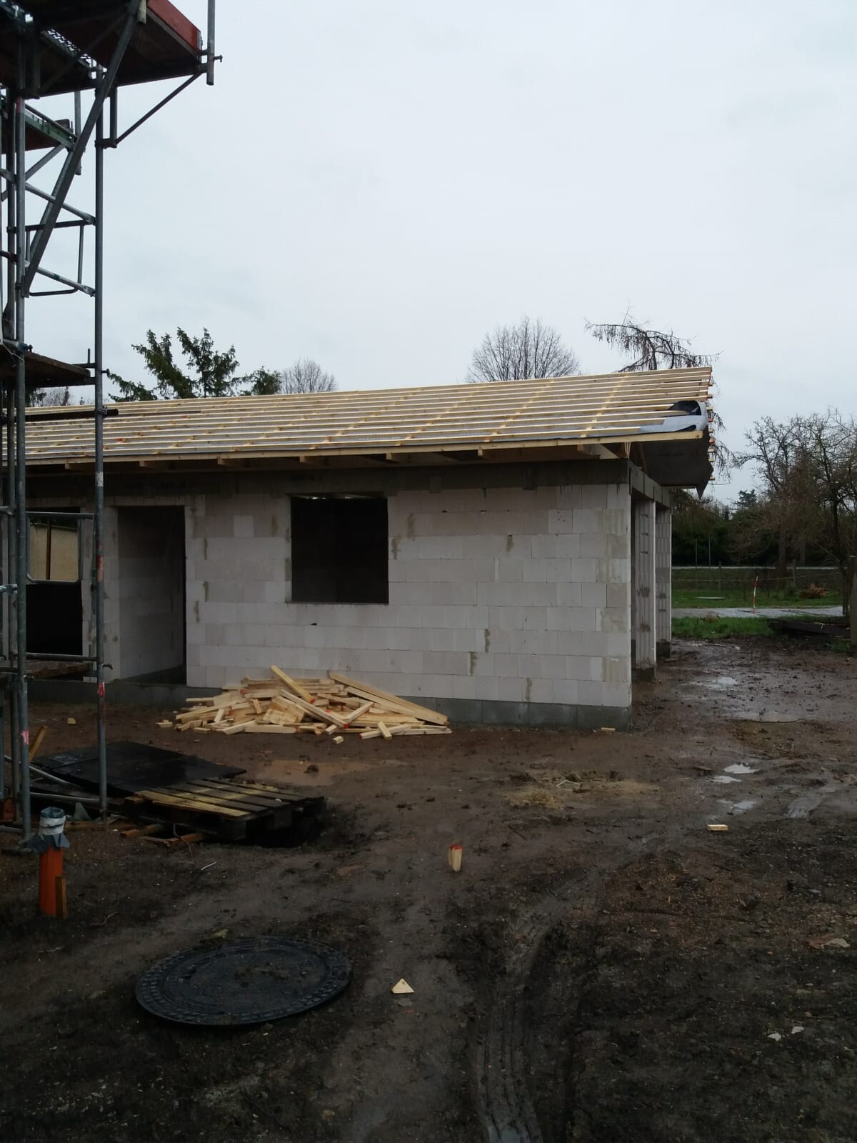 Small building made of concrete blocks, wooden roof structure, wood scraps on the ground, scaffold on the left.