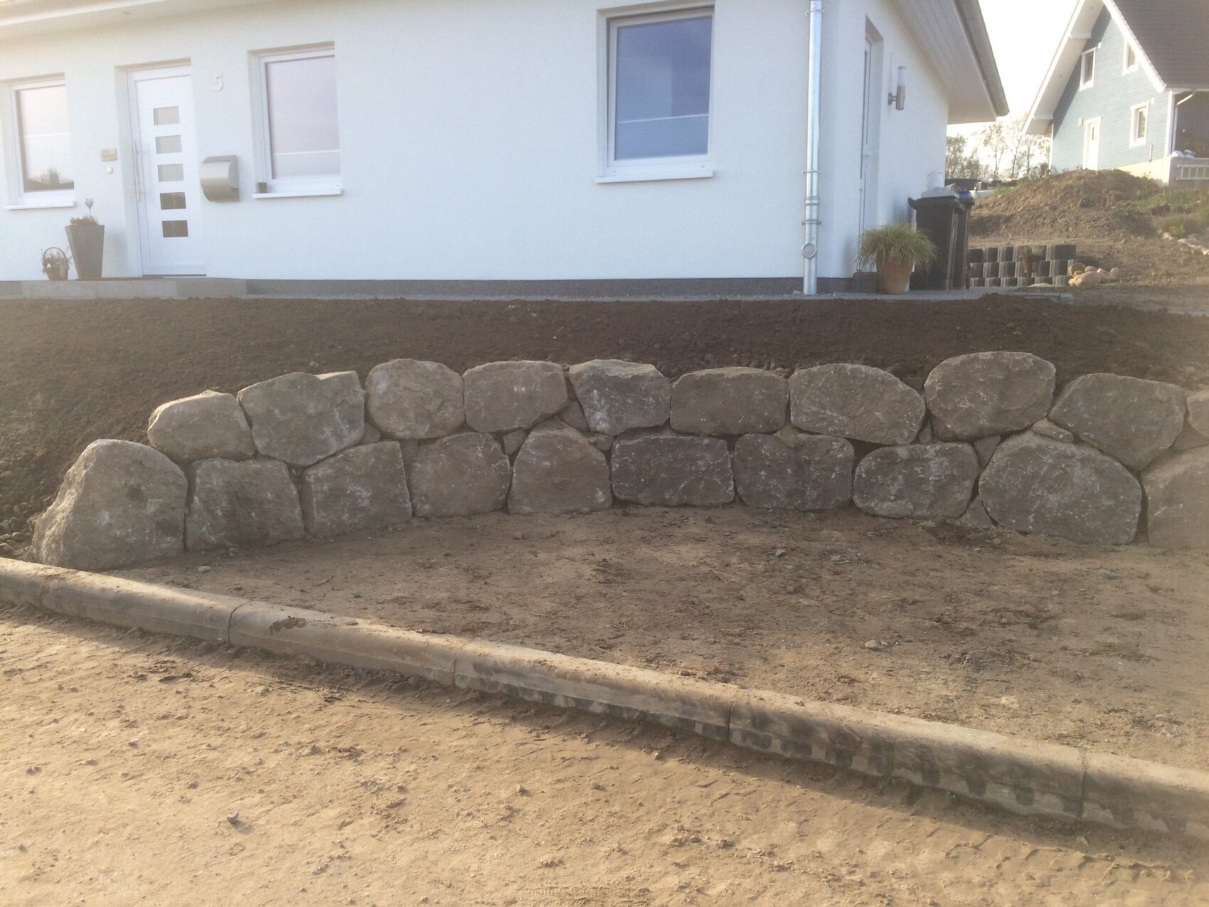 Gartenvorplatz mit einer großen Steinmauer vor dem hellen Haus und Eingang