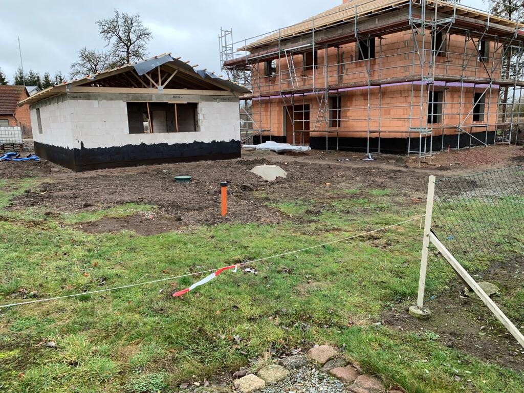 Construction site: small white block structure with black foundation waterproofing next to shell construction with scaffolding.