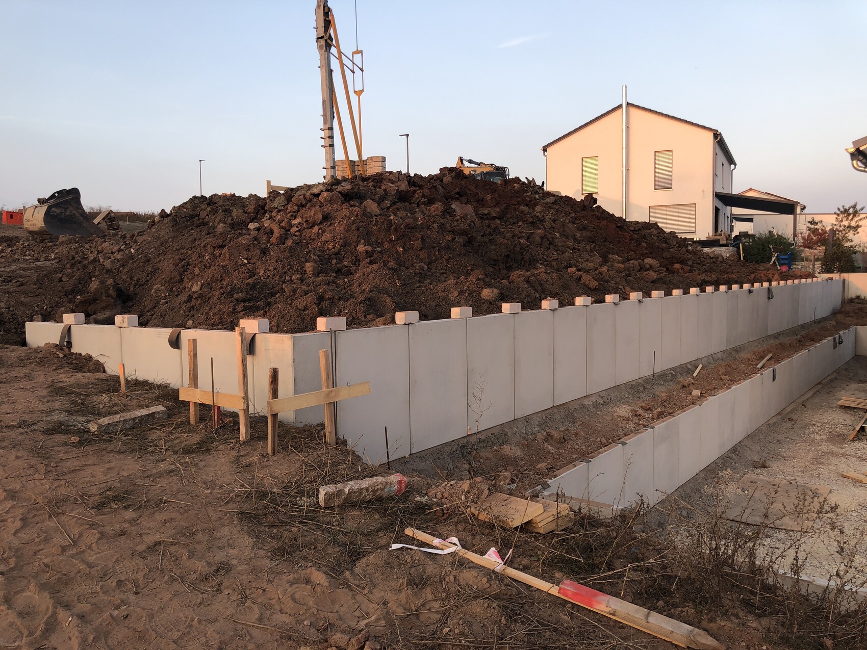 Large earth embankment behind a concrete wall; excavator arm and house in the background.