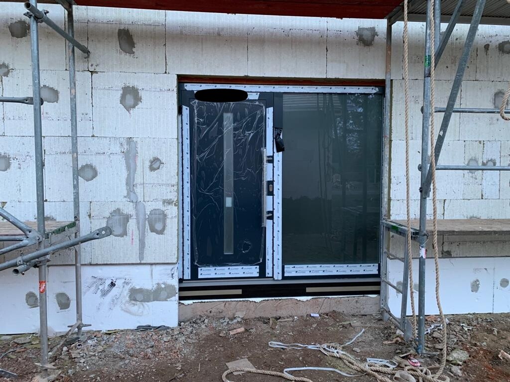 Construction site with scaffolding, unfinished facade made of insulation boards, and a glass door at the entrance.