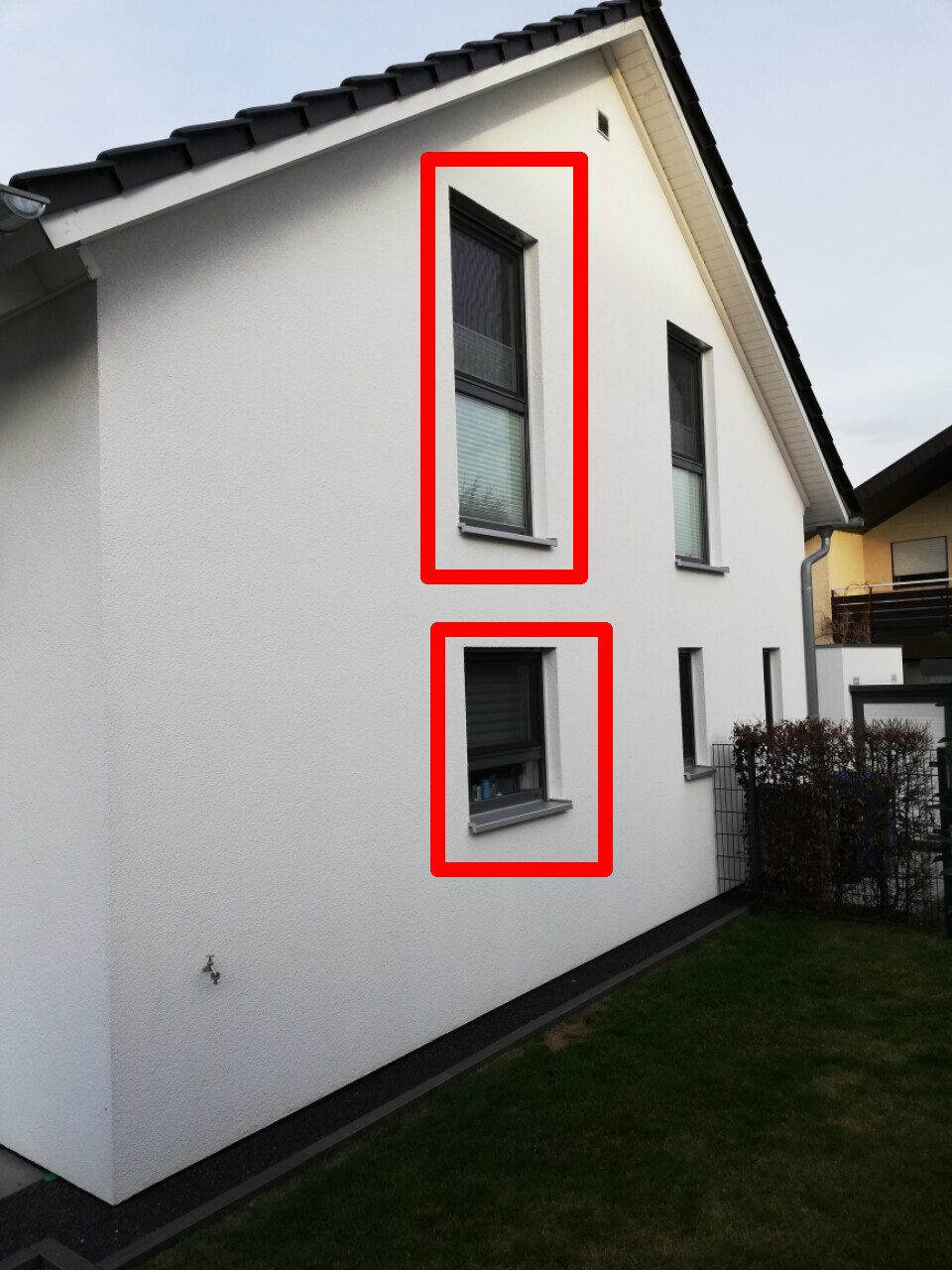 Weiße Hausfassade mit zwei Fenstern, eines im Obergeschoss und eines im Erdgeschoss.