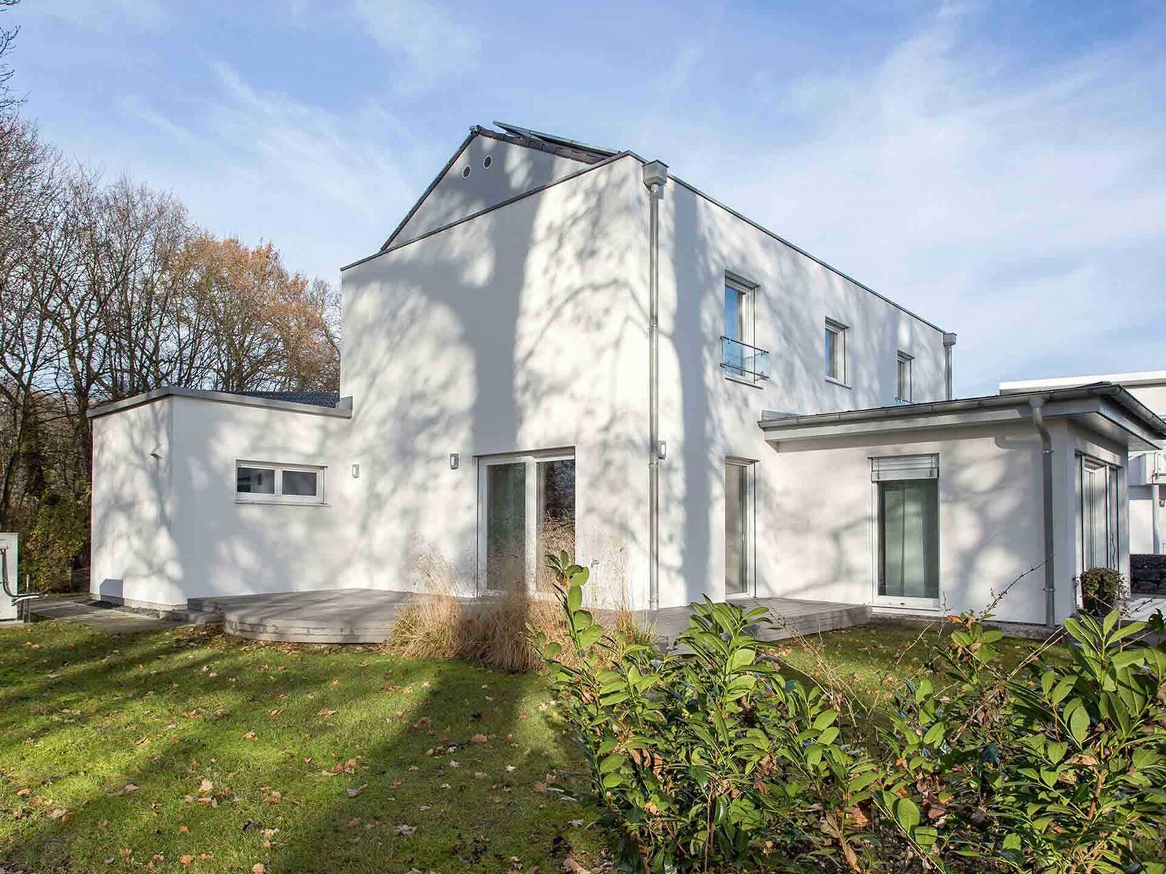 Modern single-family house in white with terrace, large windows, garden, and shadows from trees.