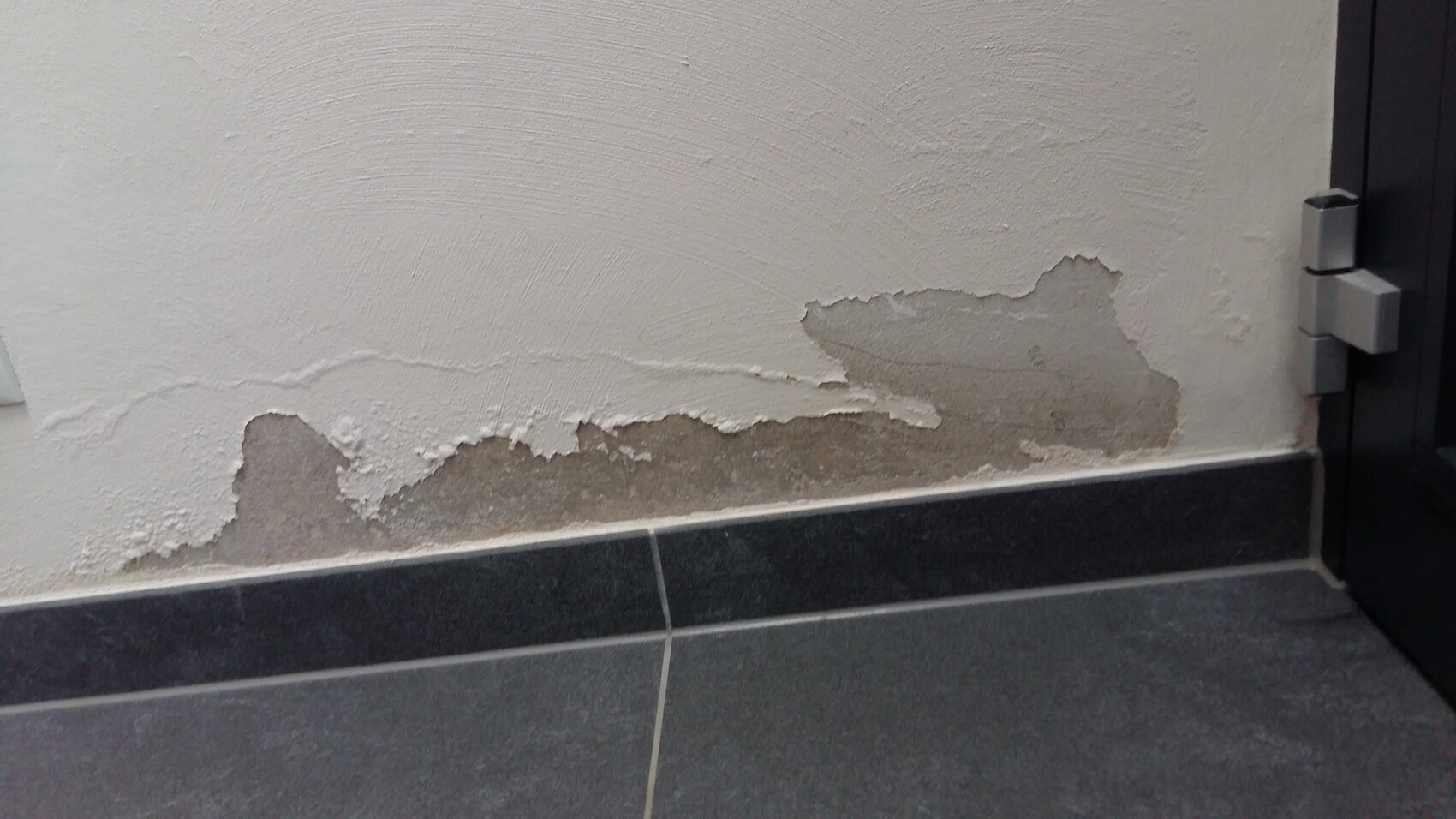 Wall with peeling plaster and exposed substrate above dark tiles; door frame on the right.