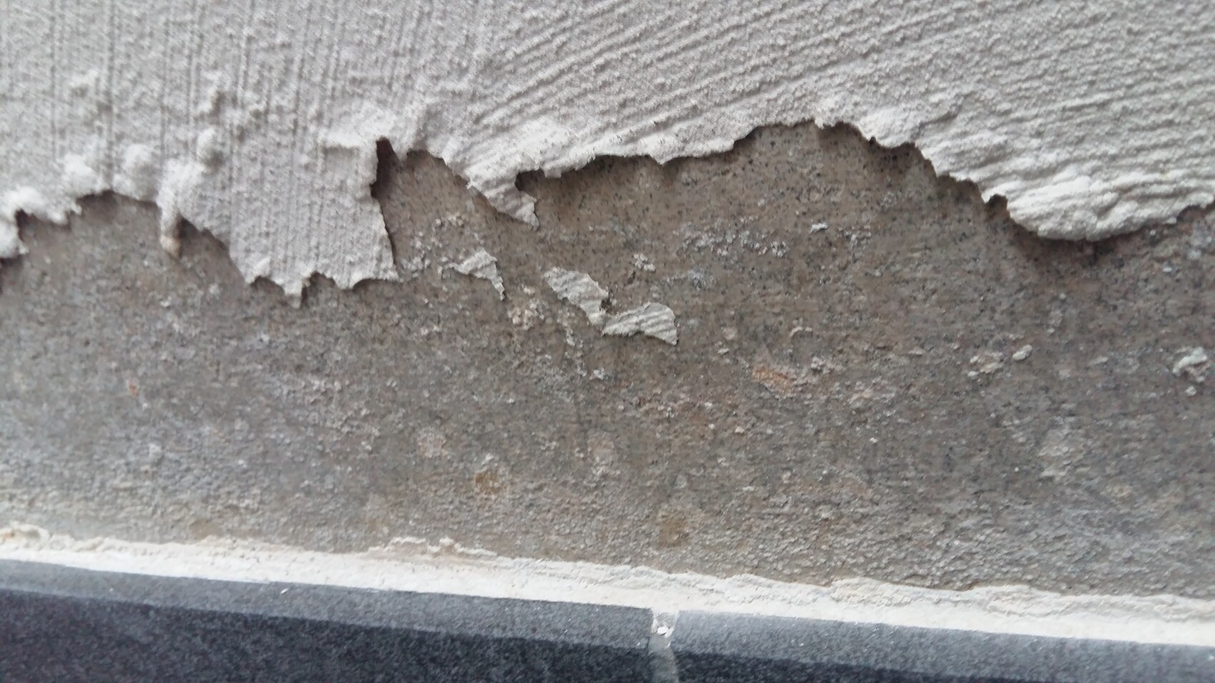 Wall with peeling plaster; rough concrete substrate and base edge visible underneath.