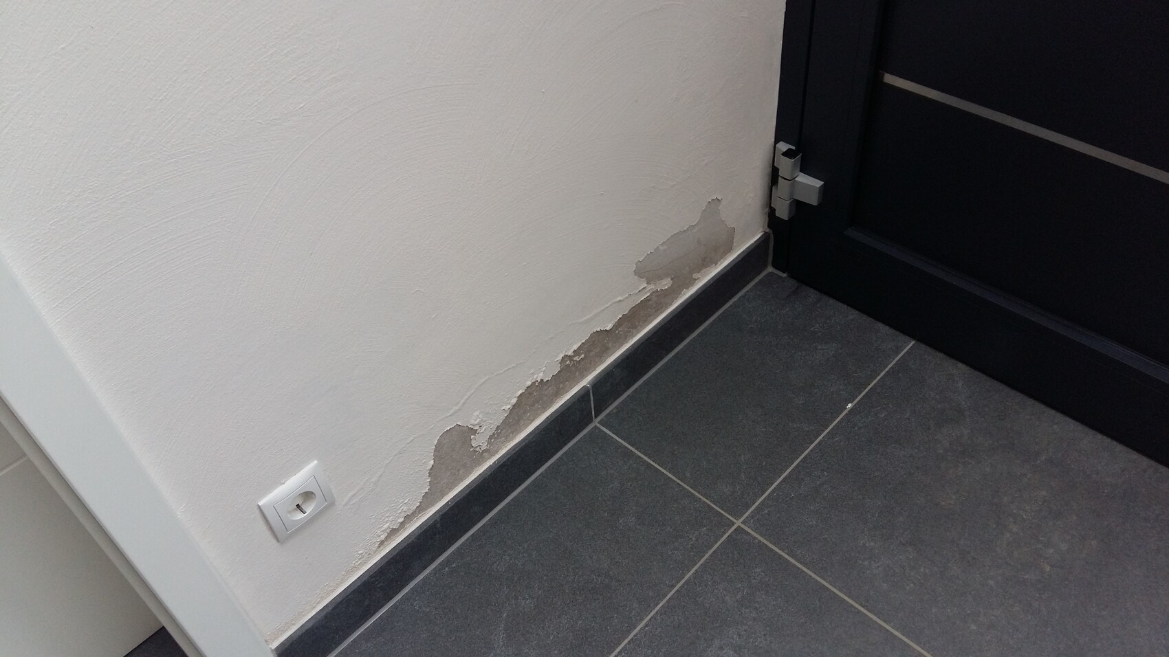 White wall with chipped plaster at the lower edge next to a black door; dark tile floor.