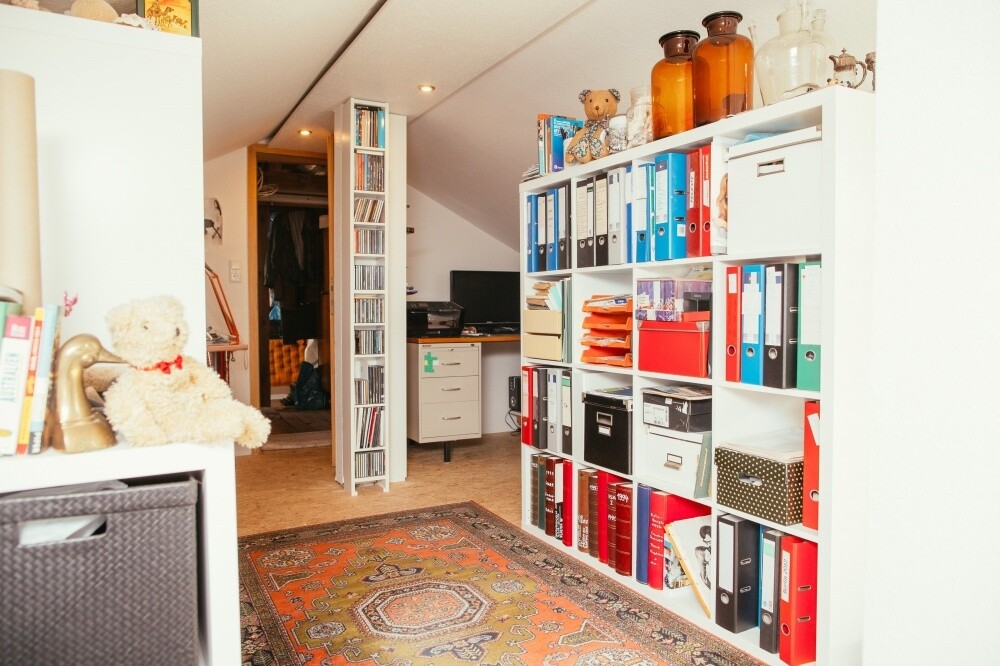 Helles Arbeitszimmer mit weißen Regalen voller Ordner, Schreibtisch und Teppichboden