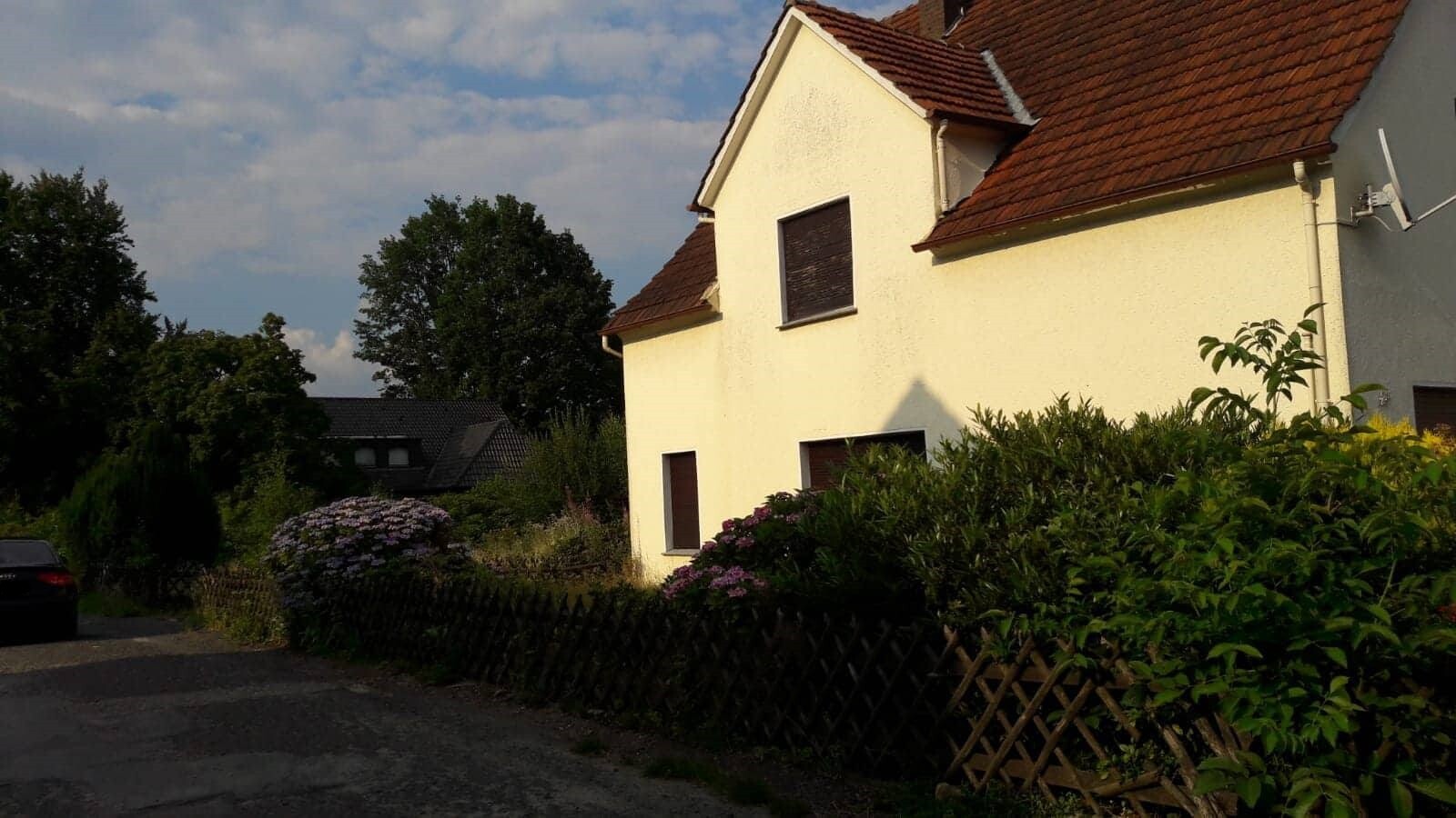 Gelbes Einfamilienhaus mit rotem Ziegeldach, Gartenzaun, Hydrangea-Sträucher und davor geparktes Auto.