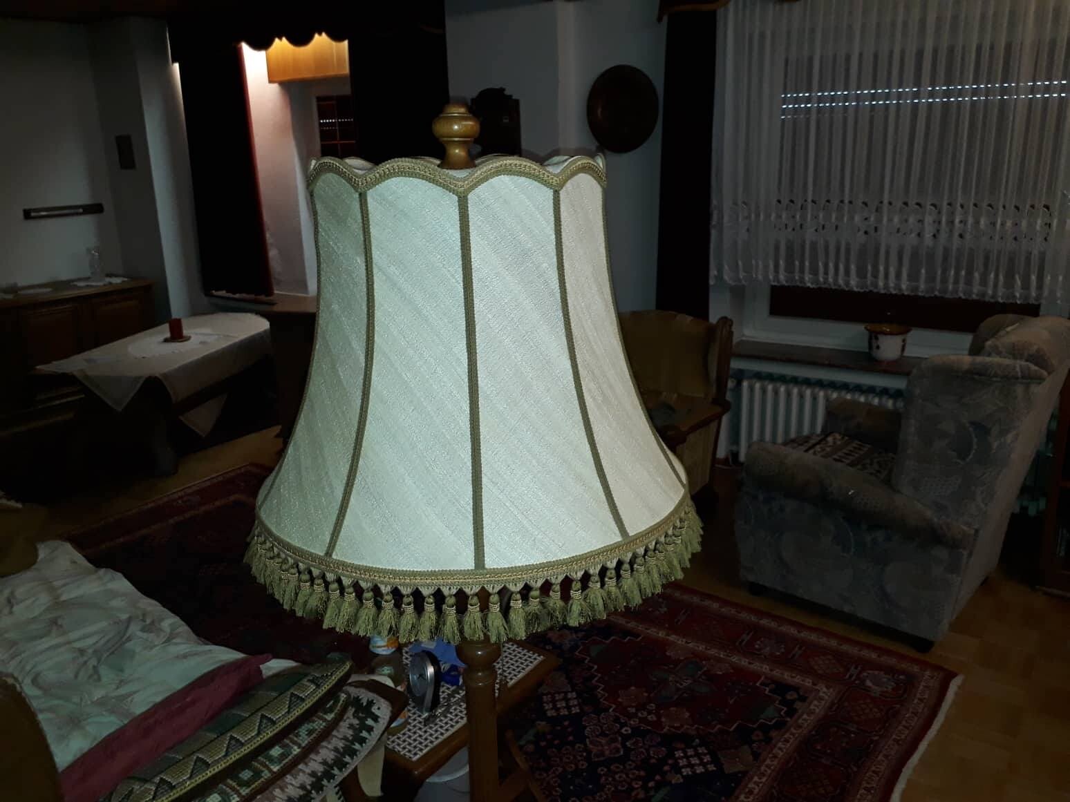 Große cremefarbene Stofflampe mit goldener Zierkante und Fransen im Wohnzimmer, Sofa im Hintergrund.