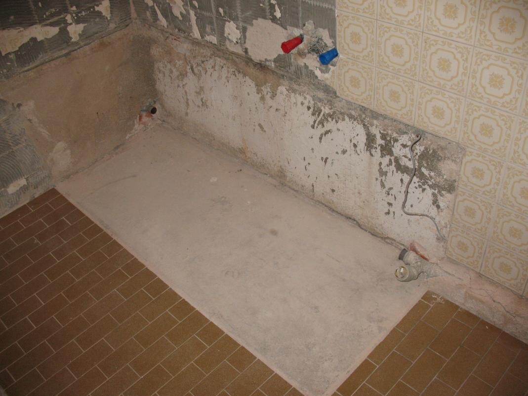 Unfertiges Badezimmer mit freiliegenden Rohren, feuchter Wand und rohem Boden