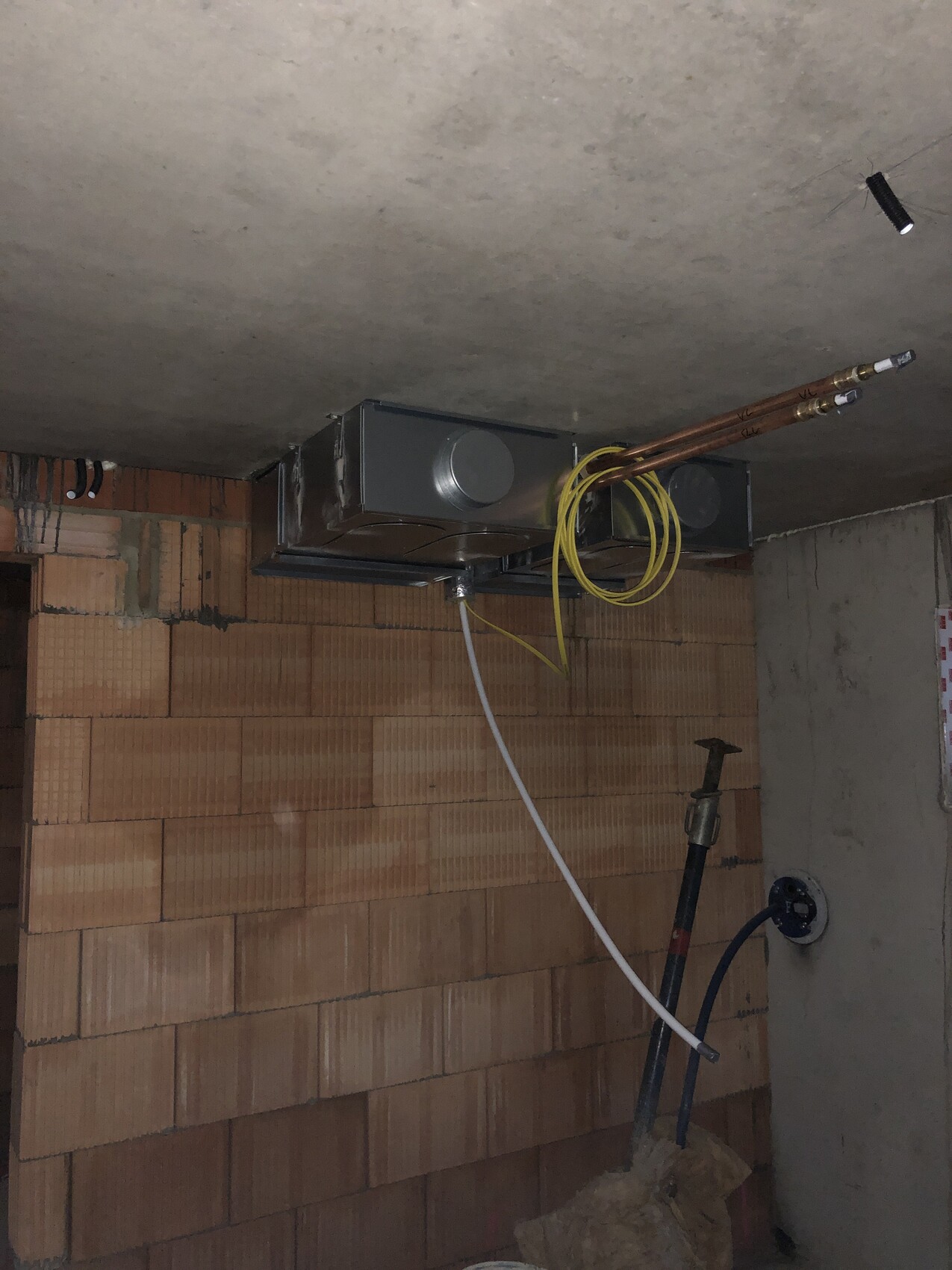Basement ceiling: ventilation unit with two copper pipes, yellow cables attached to brick wall.