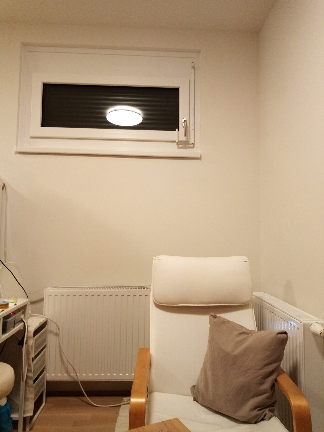 White armchair with brown cushion in front of radiator and window high on the wall.