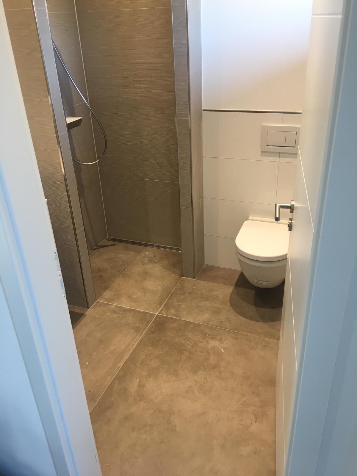 Small bathroom: shower on the left, wall-mounted toilet on the right, gray floor and brown wall tiles.