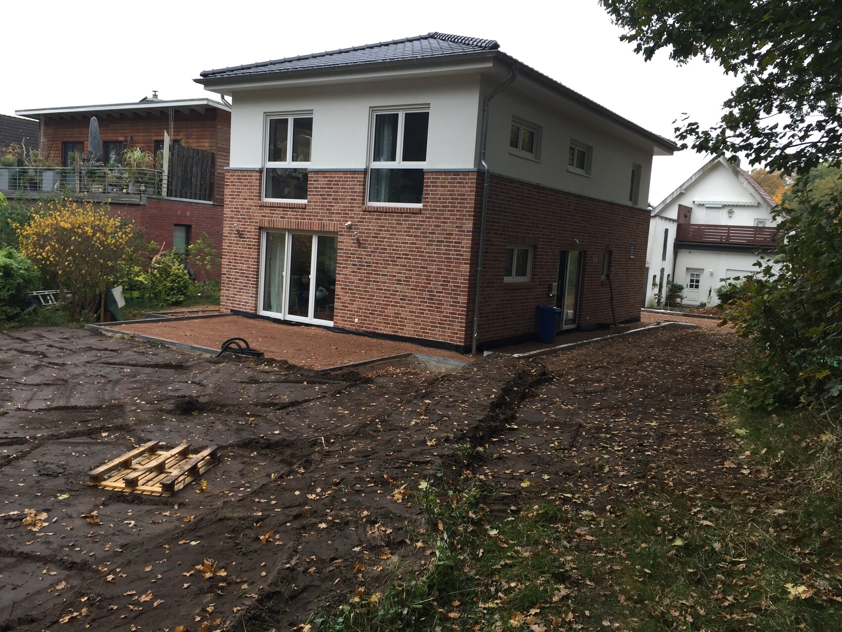 Two-story brick house; garden construction site with earthworks and pallets.