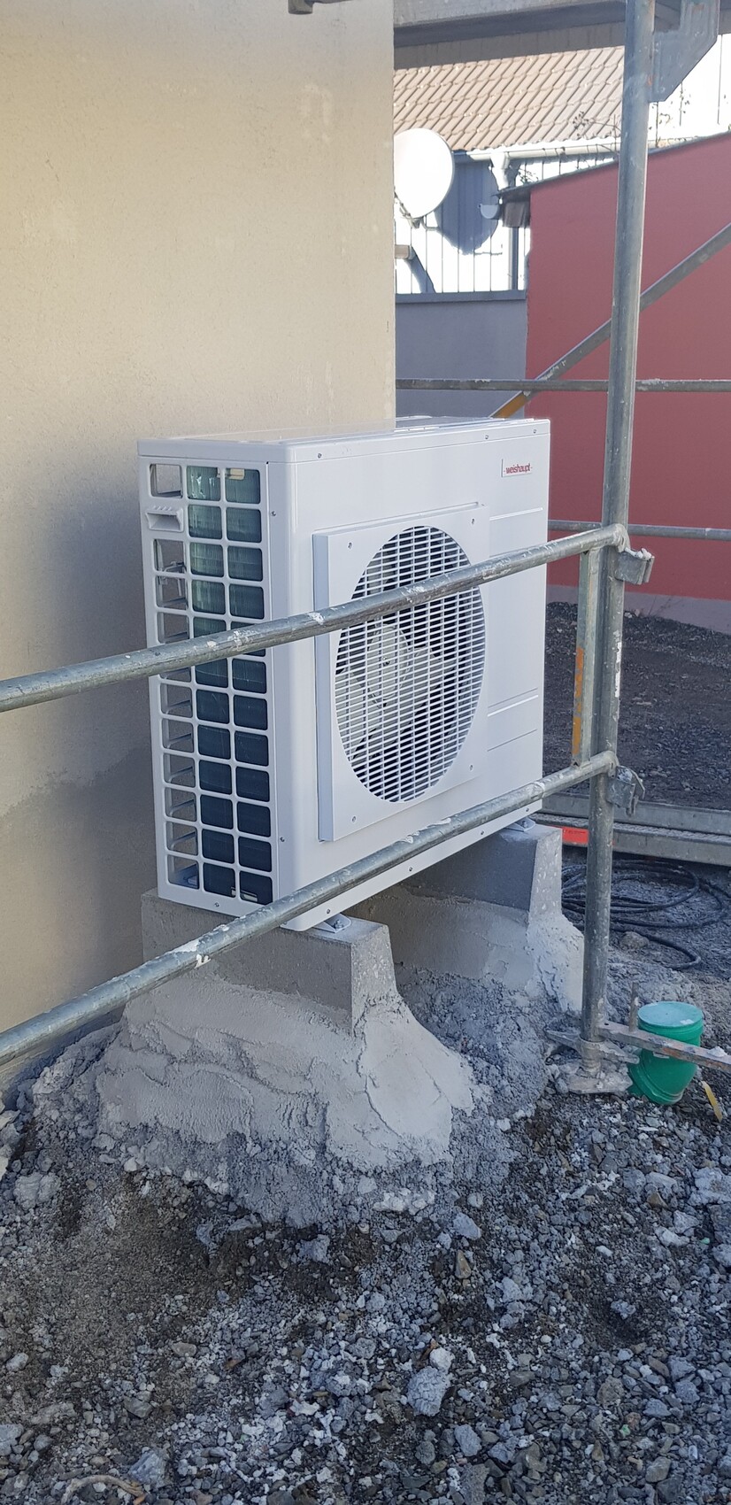 Außen-Kondensator einer Split-Klimaanlage an einer Wand, von Gerüststangen umgeben, Kiesboden darunter