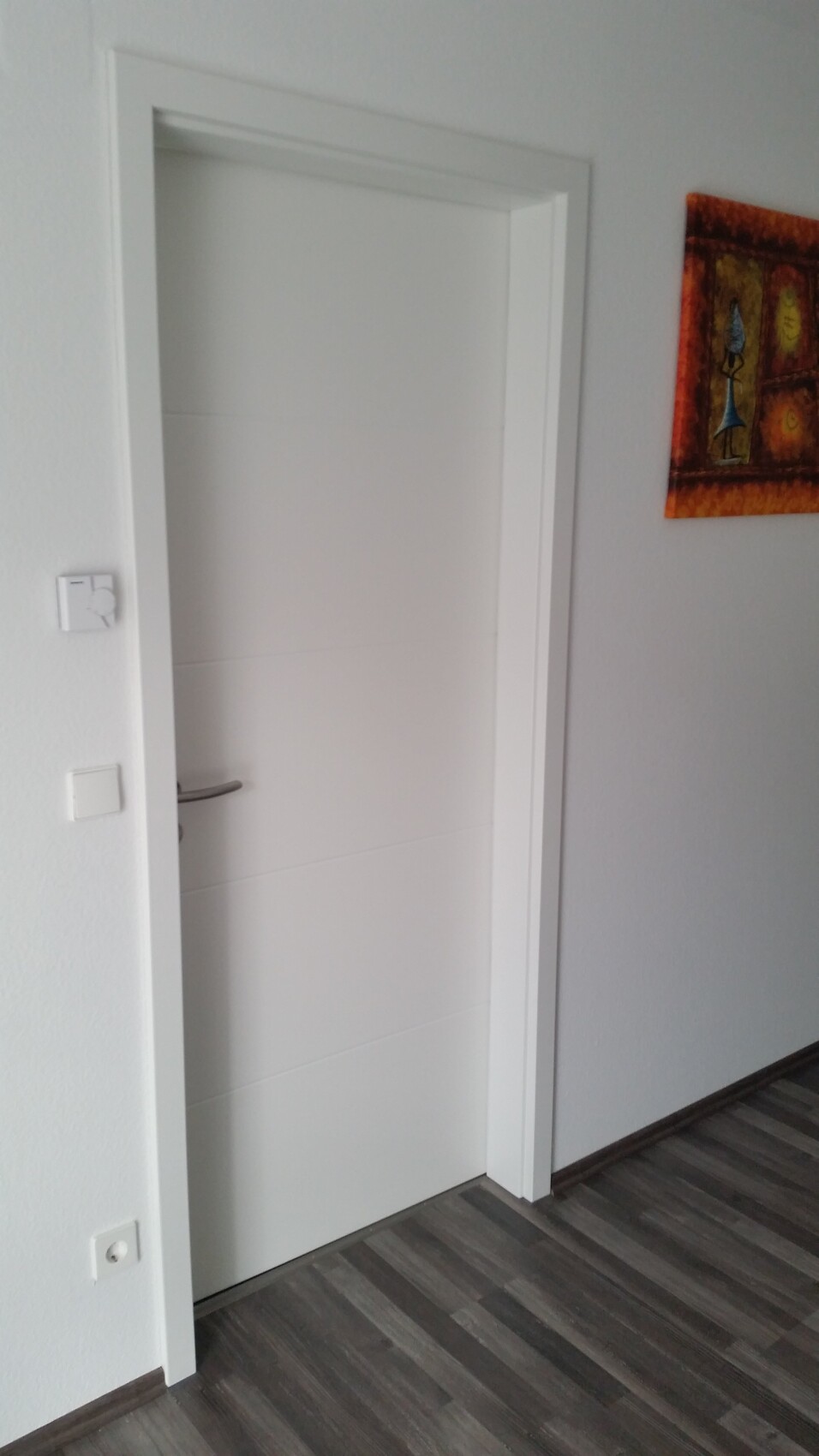 White interior door with handle in the hallway, colorful painting to the right; dark laminate floor.