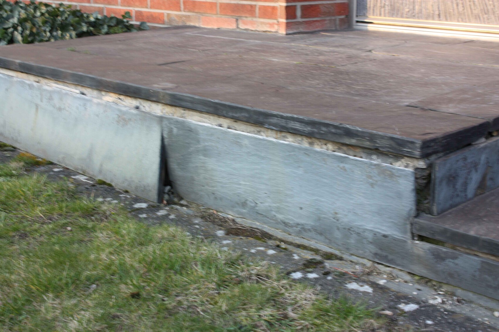 Unfinished terrace with concrete foundation, visible construction joint and grass in front, red brick house in the background.