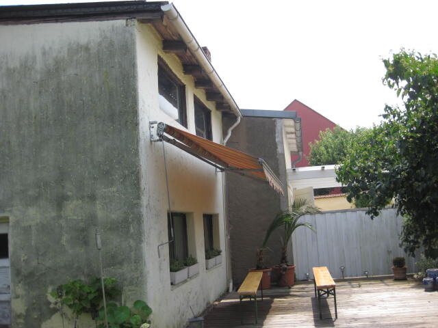 Außenansicht eines Hauses mit Holzdeck, ausziehbarem Sonnenschutz, Bänken und Topfpflanzen.