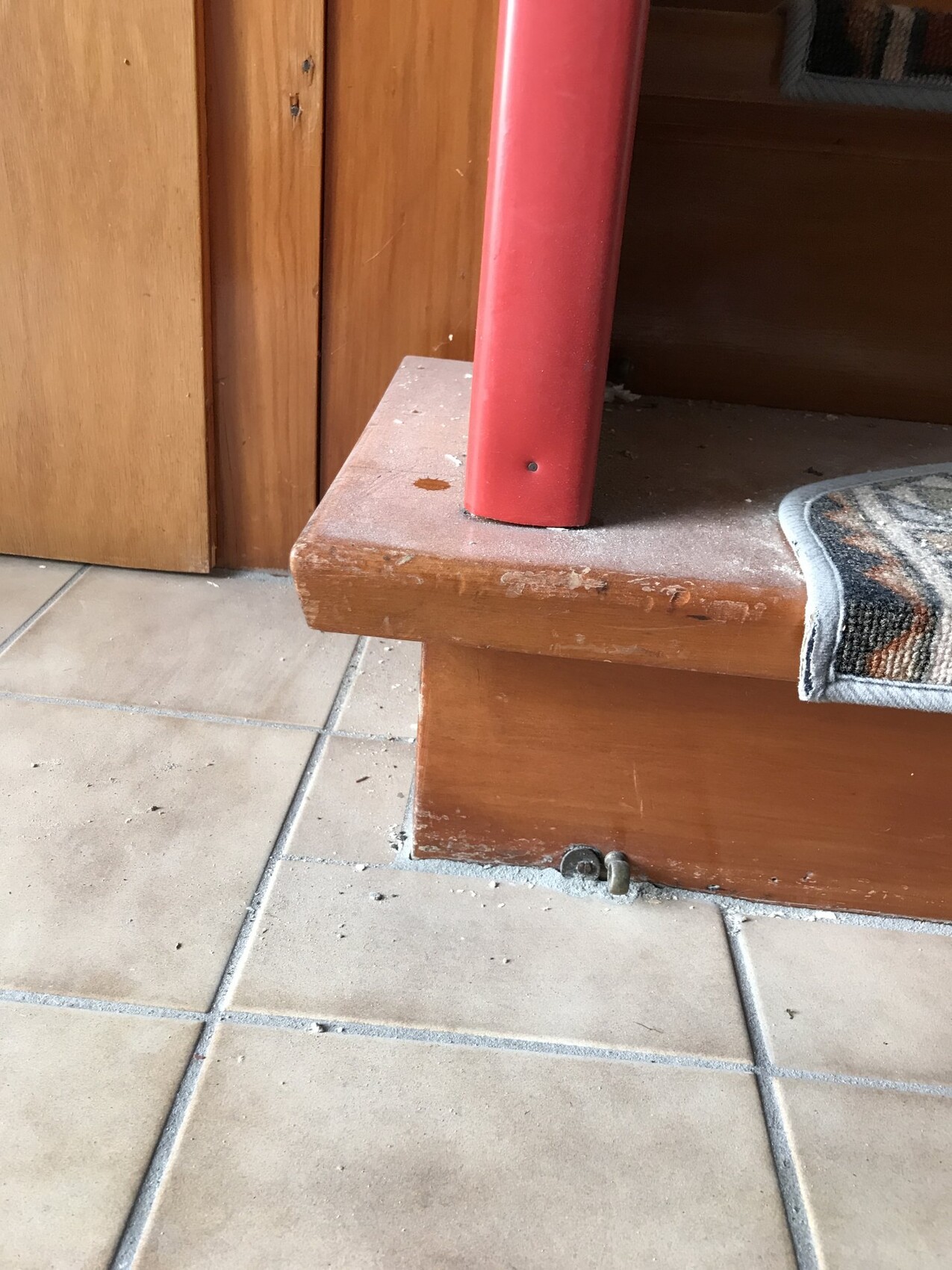 Close-up of a wooden bench with red support on tiled floor, dust and carpet edge visible.