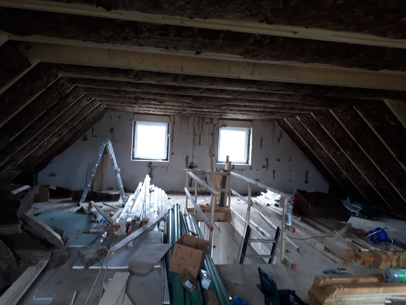 Attic construction: exposed roof beams, wood, ladder, and building materials in front of two windows.