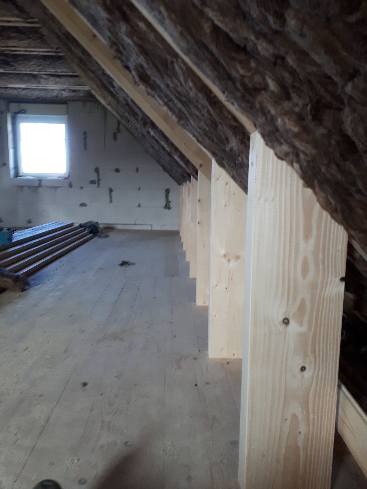 Attic construction site: visible wooden posts, roof insulation, wooden floor, window at the end.