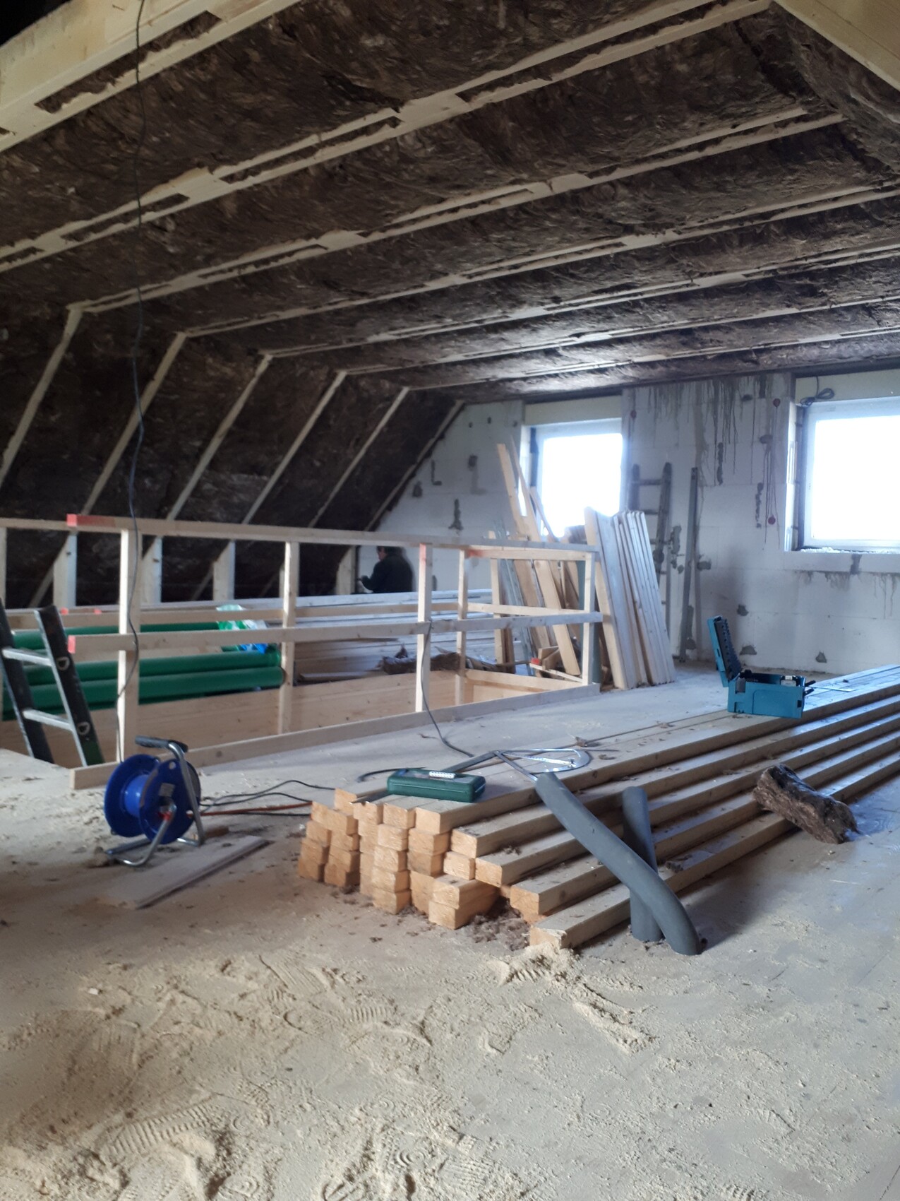 Attic construction site: stacked wooden slats, beams, tools, visible worker.