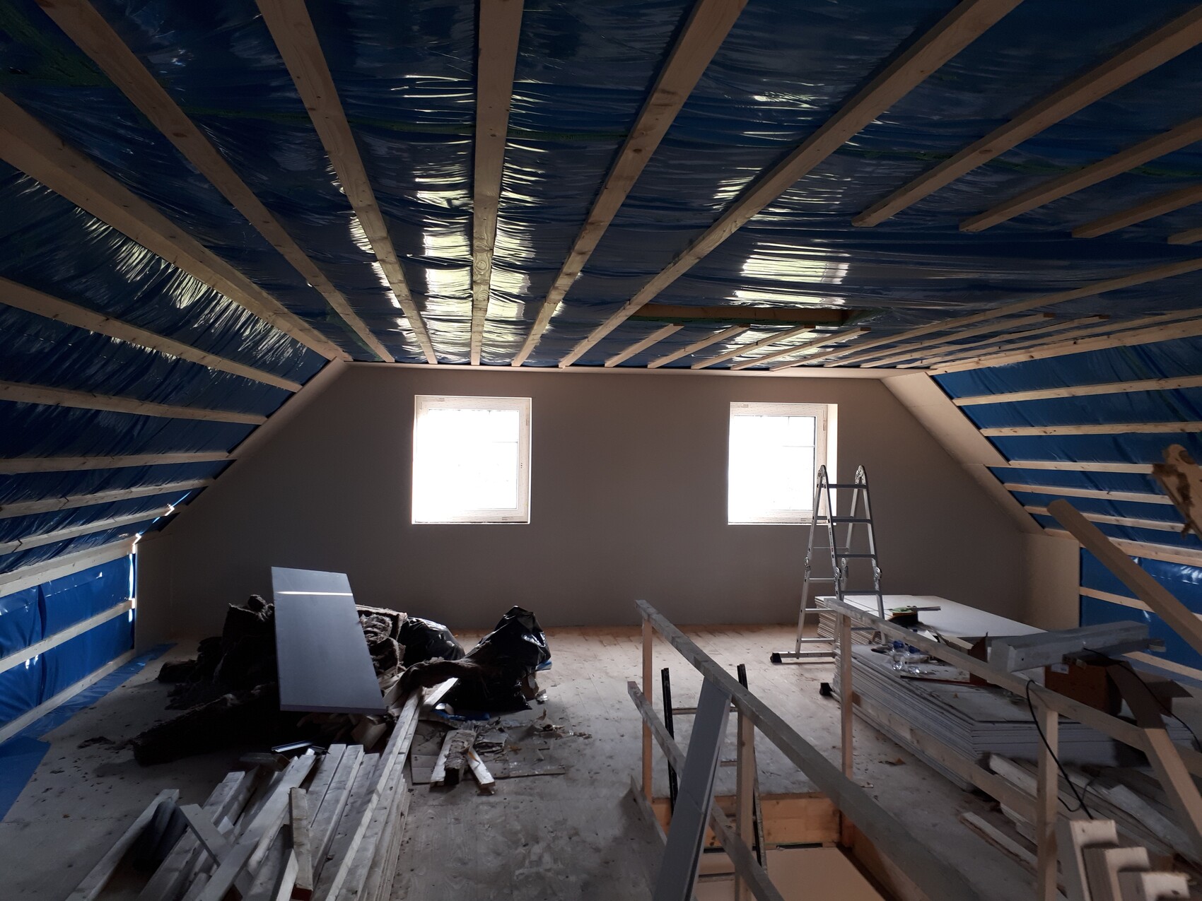 Shell attic with blue insulation membrane, two windows, wooden beams, ladder, and construction lumber.