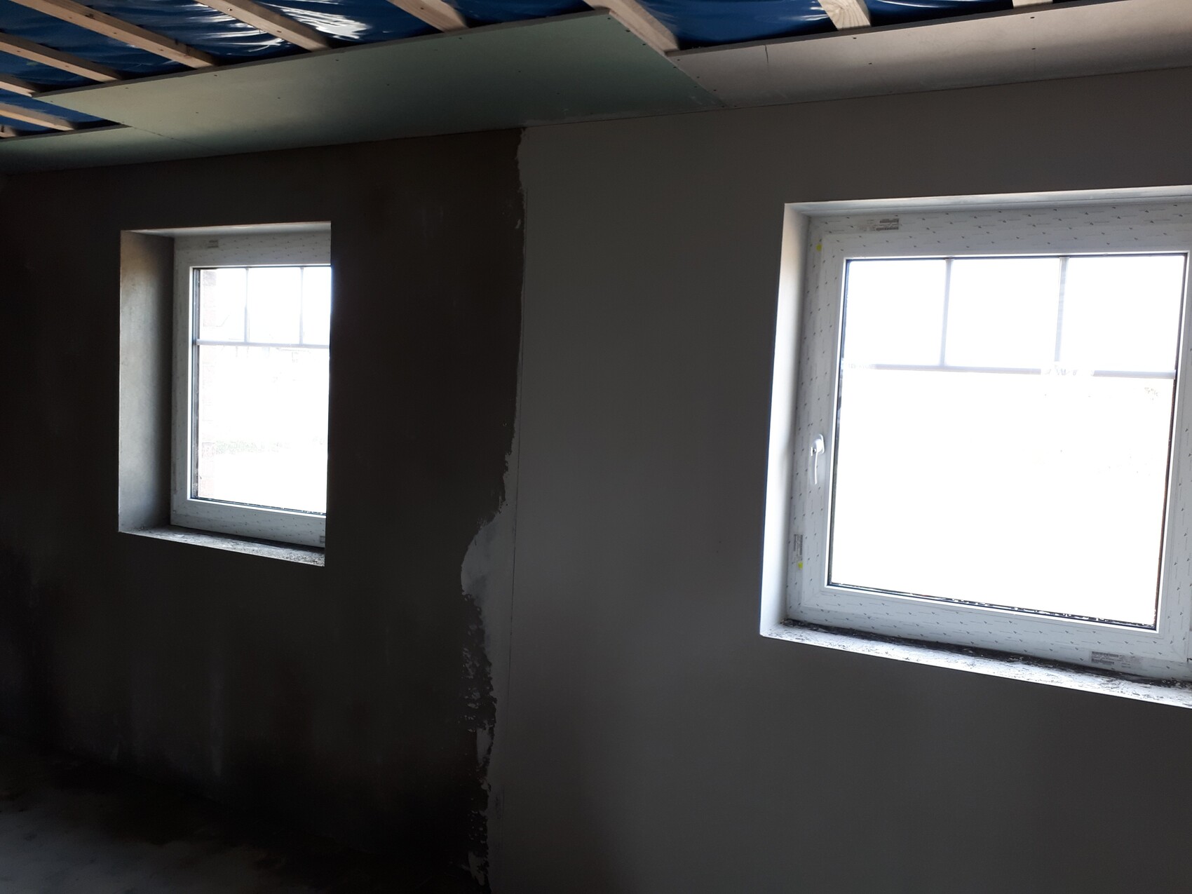 Interior space under construction: two windows in the wall; left smaller, right larger; open ceiling.
