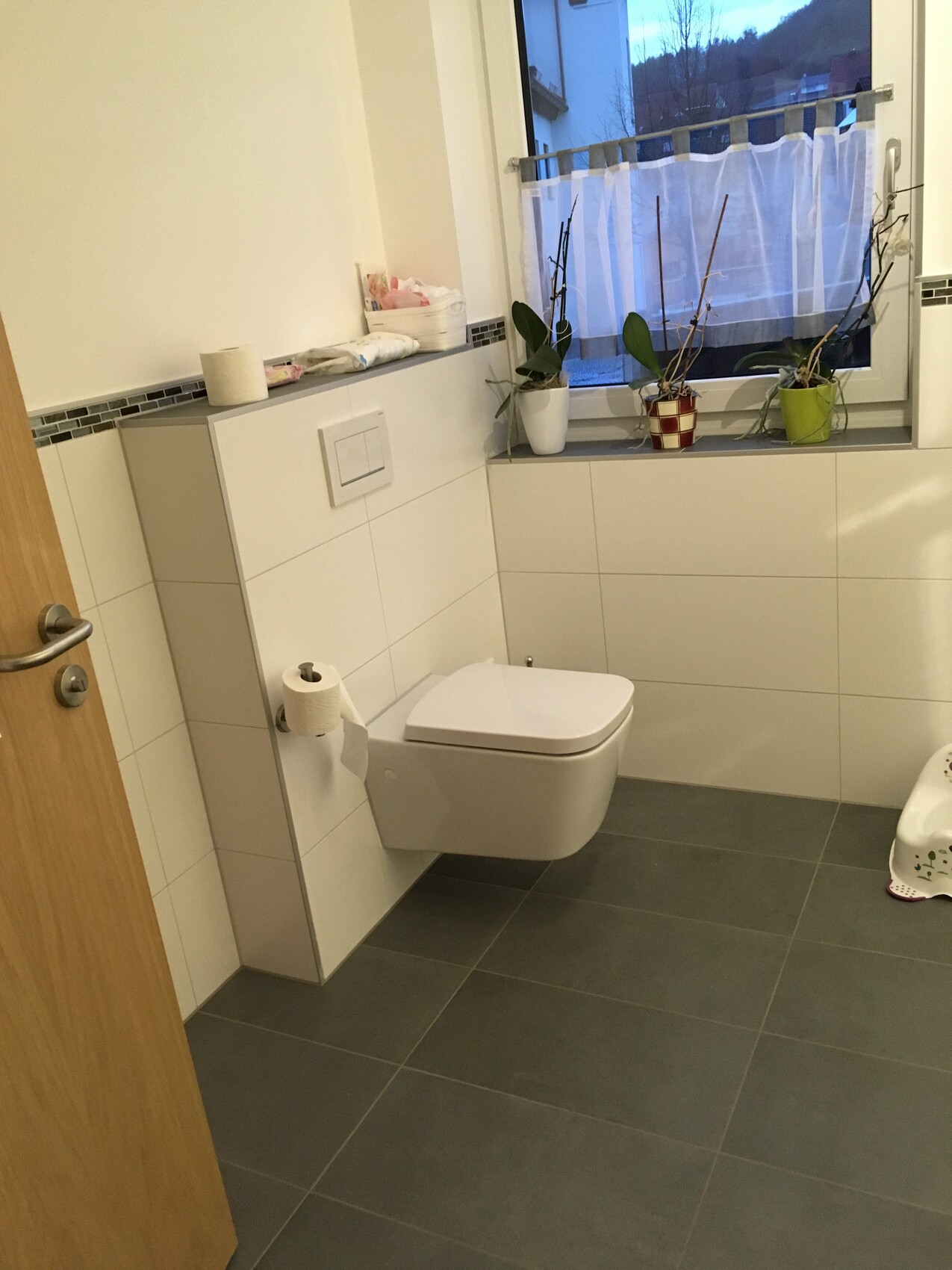 Modern bathroom with wall-hung toilet, white tiles, and window plants.