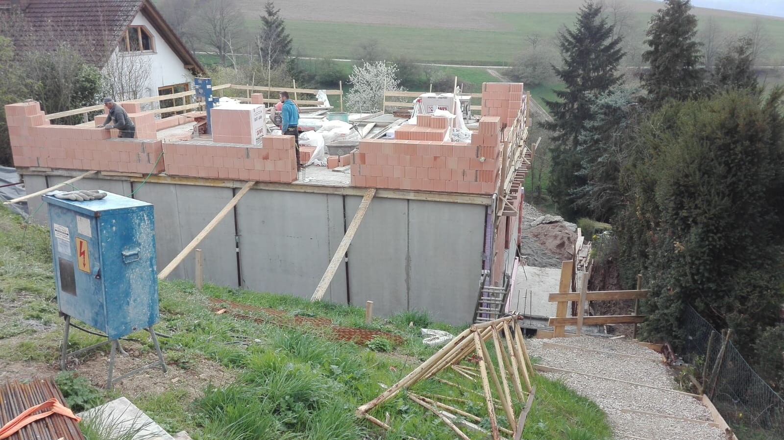 Shell construction: brick walls above foundation, construction workers working on site in a green area.