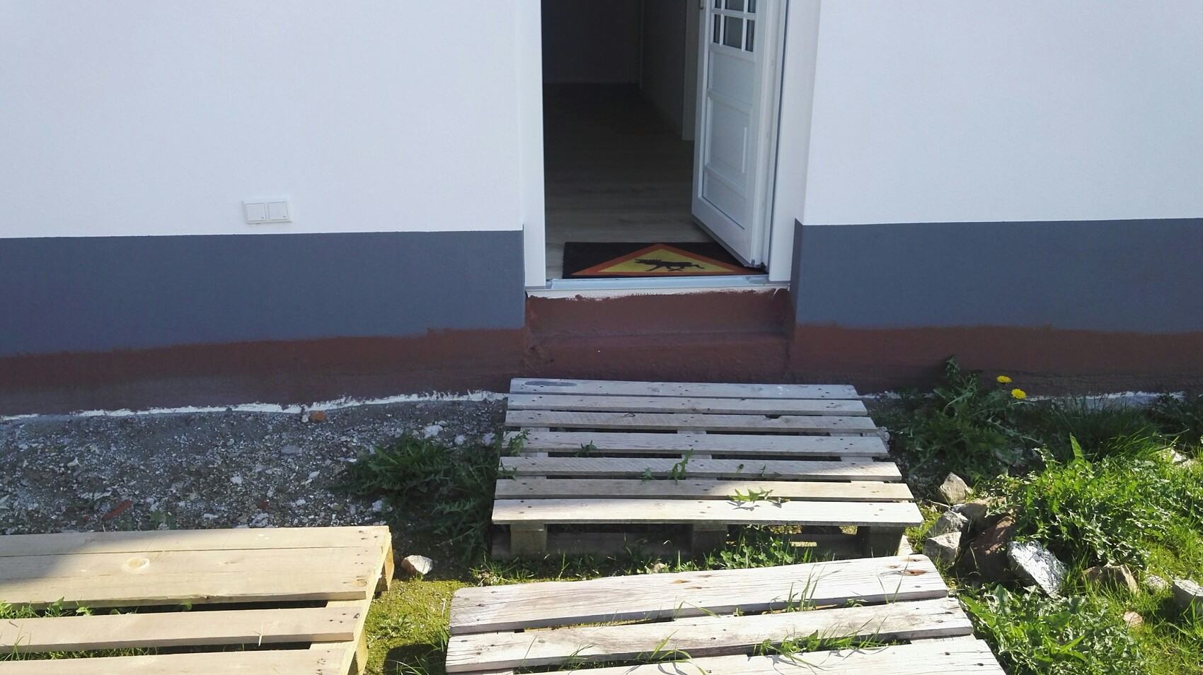 Open front door of a white house; yellow warning sign on the doormat, pallet path to the entrance.
