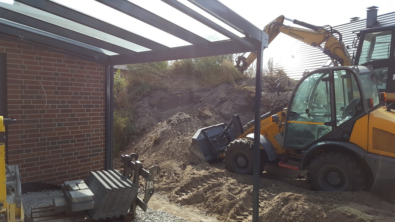 Yellow excavator at a steel frame structure in front of a red brick wall, sandy soil.