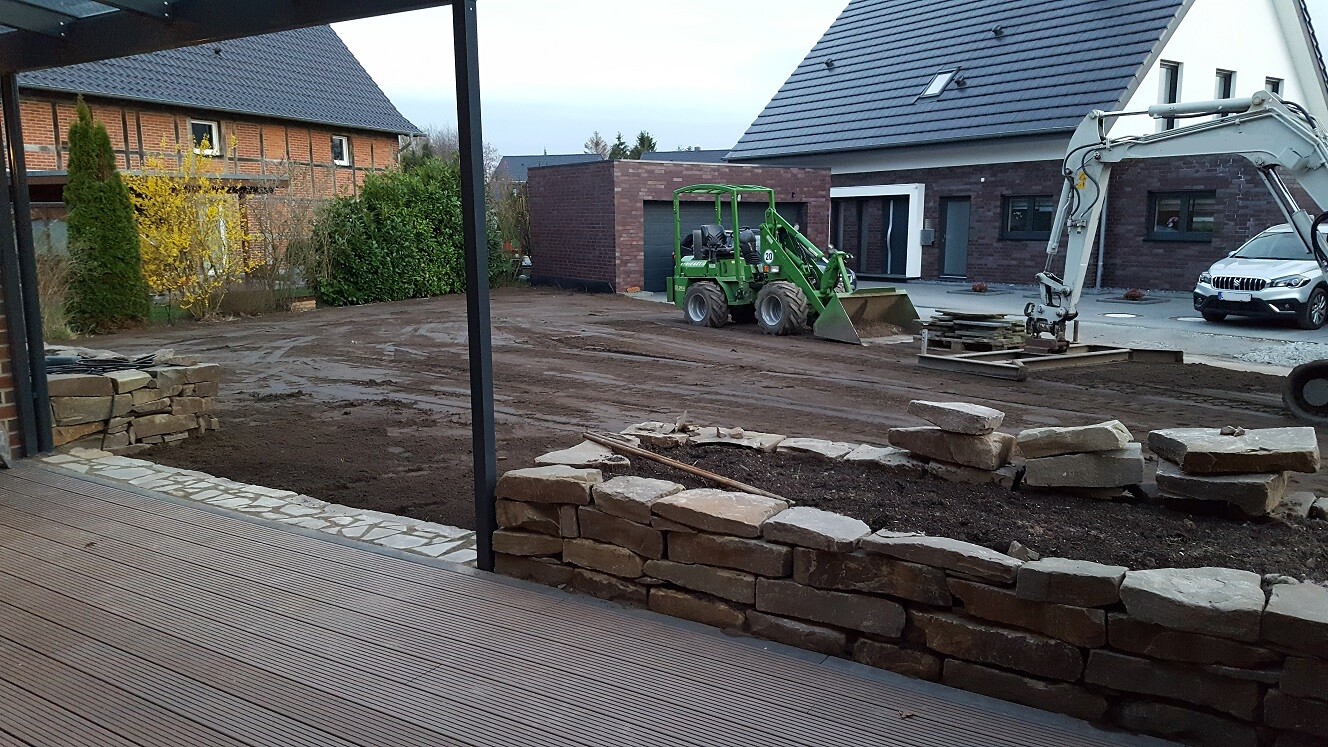 Construction site with stone wall, green compact loader, mini excavator, wooden deck, and house in the background.