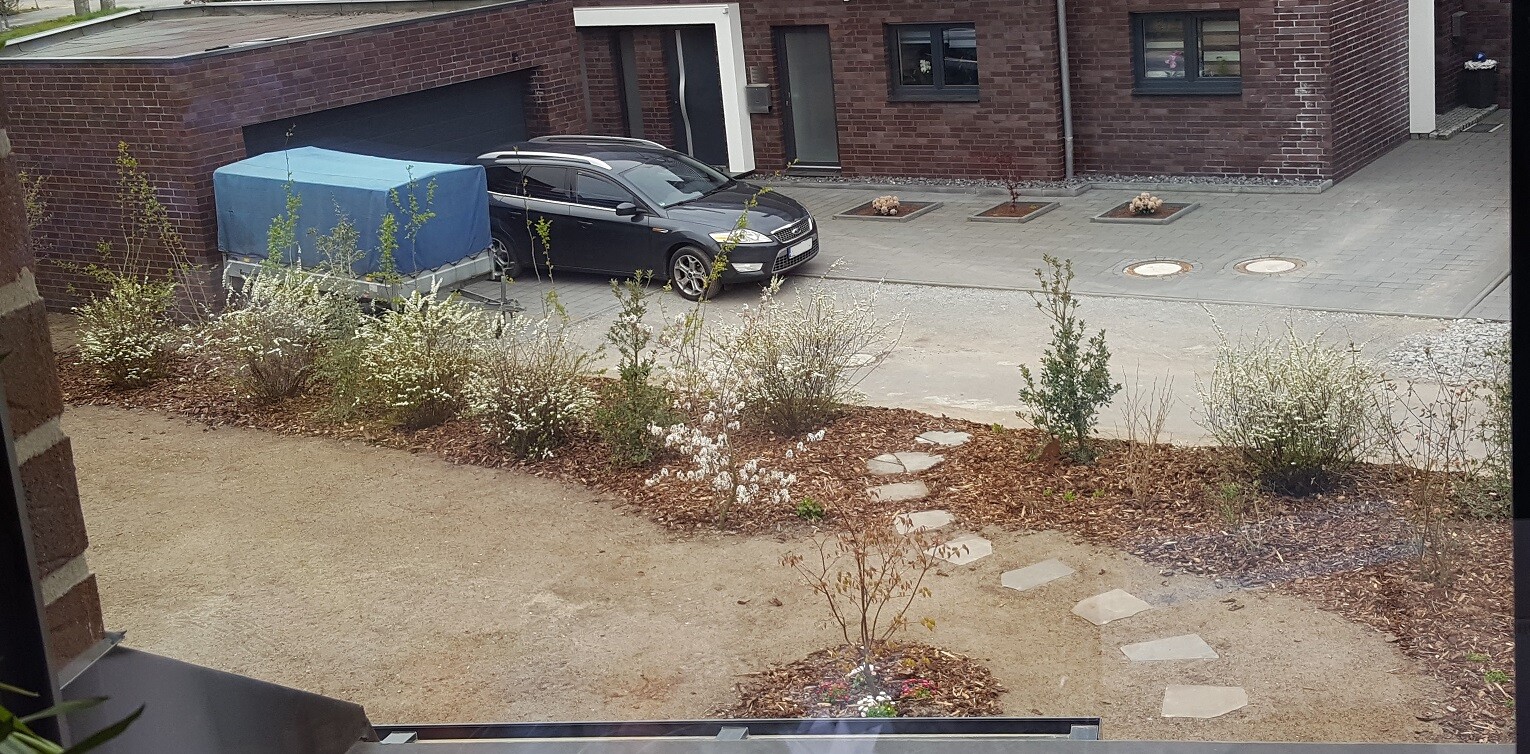 Black car in front of the garage of a brick house; blue covered trailer, gravel path, and bushes.