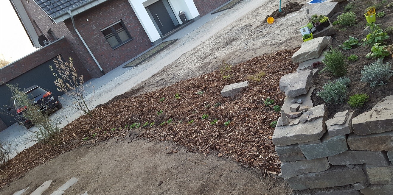 Front garden in front of a modern house: rubble stone retaining wall on the right, mulched bed, and plants.