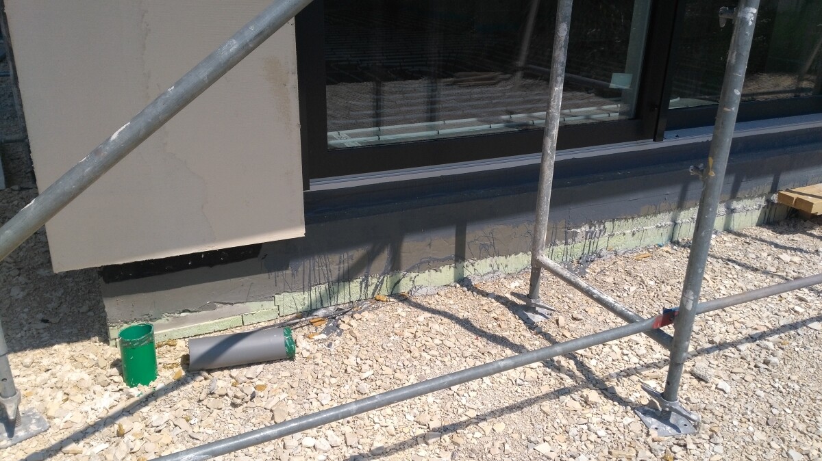 Construction site at building: scaffold pipes, gravel ground, green paint can next to window.