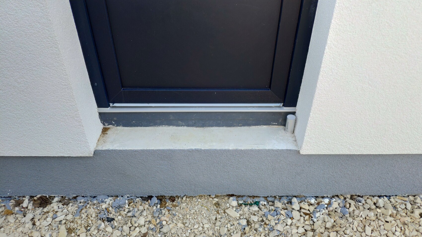 Dark door with dark frame in front of light exterior wall, front threshold, gravel ground in front, small white pipe to the right.