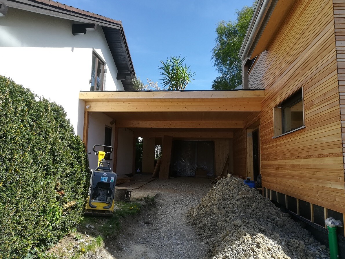 Construction site with wooden-clad extension, gravel pile, construction equipment, green hedge on the left, blue sky.
