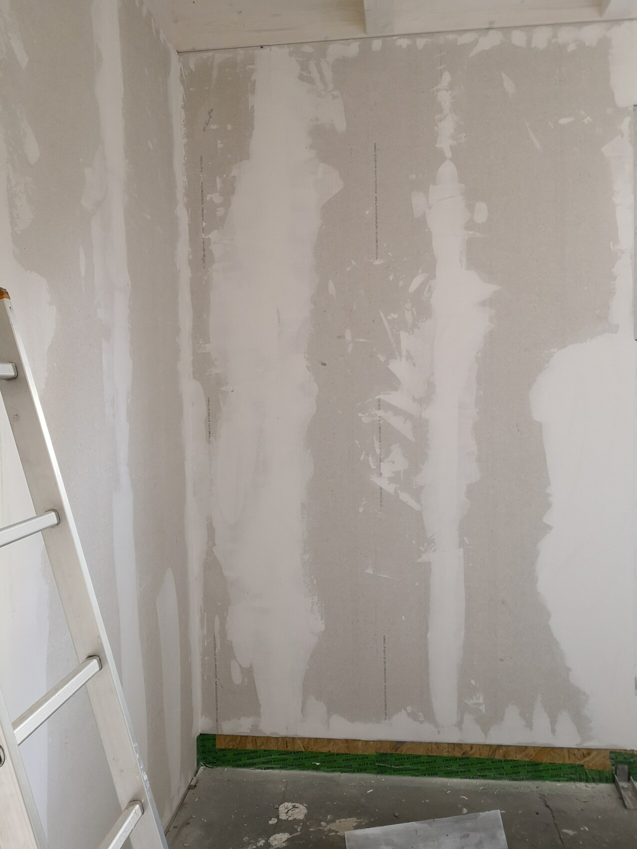 Interior under construction: skim-coated drywall, a ladder on the left, green protective foil on the floor.