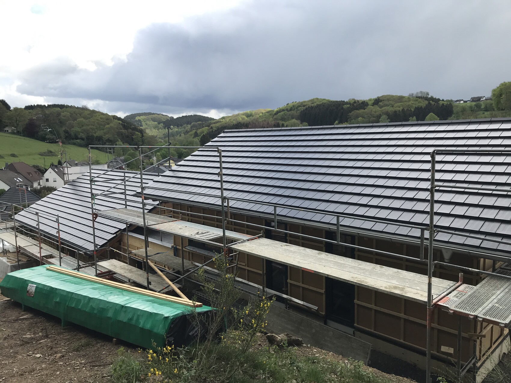 Construction site on a hillside with dark gray roof tiles, scaffolding, and green landscape.