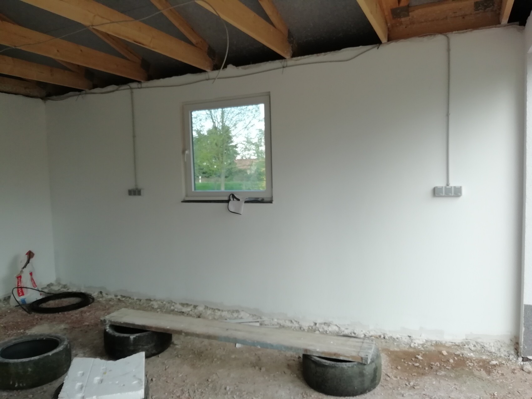 Unfinished interior: white wall, window, ceiling with wooden beams, construction tools on the floor.