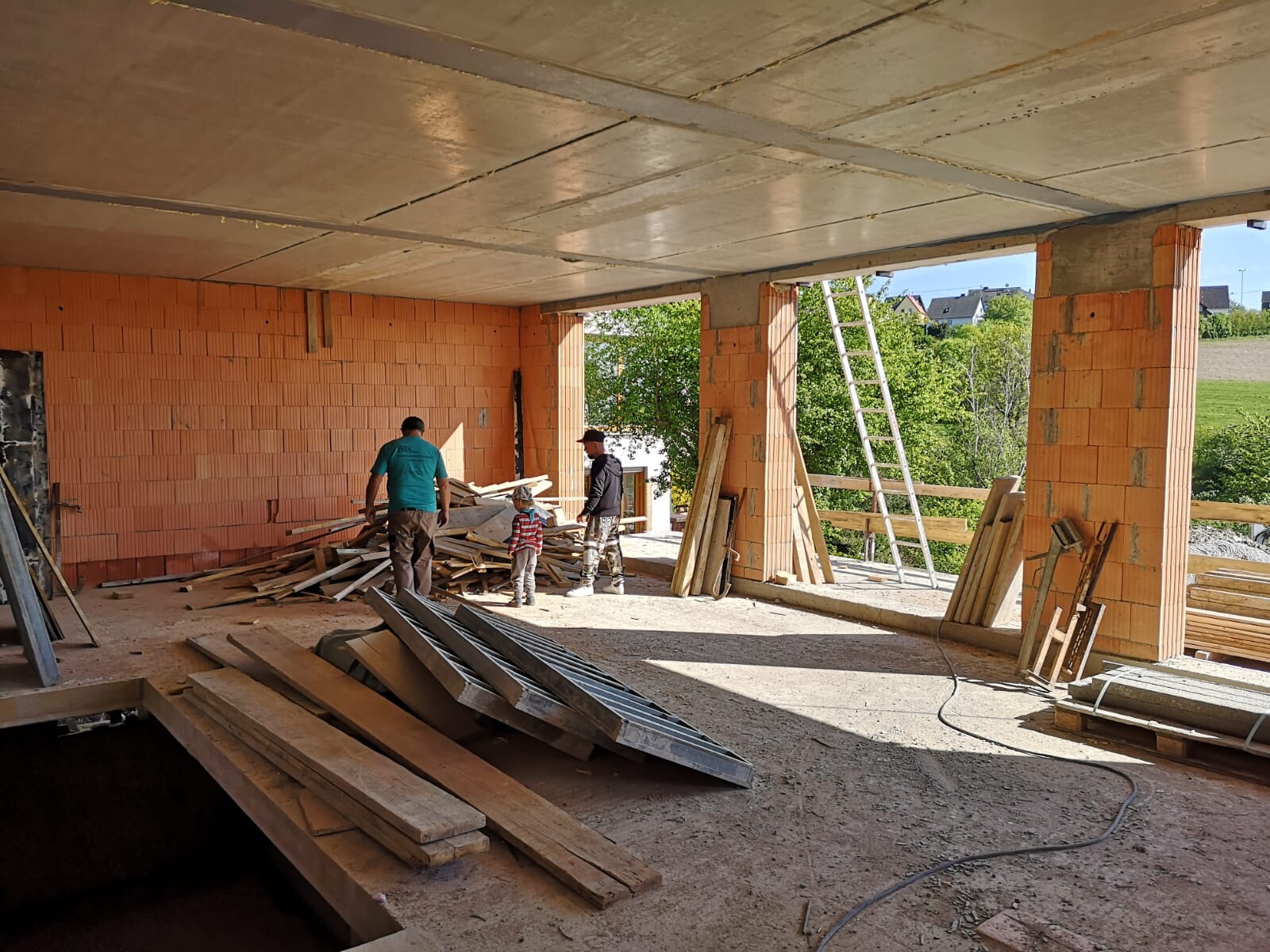 Shell construction of a brick house three people sorting wood scraps, ladder standing, outside green field