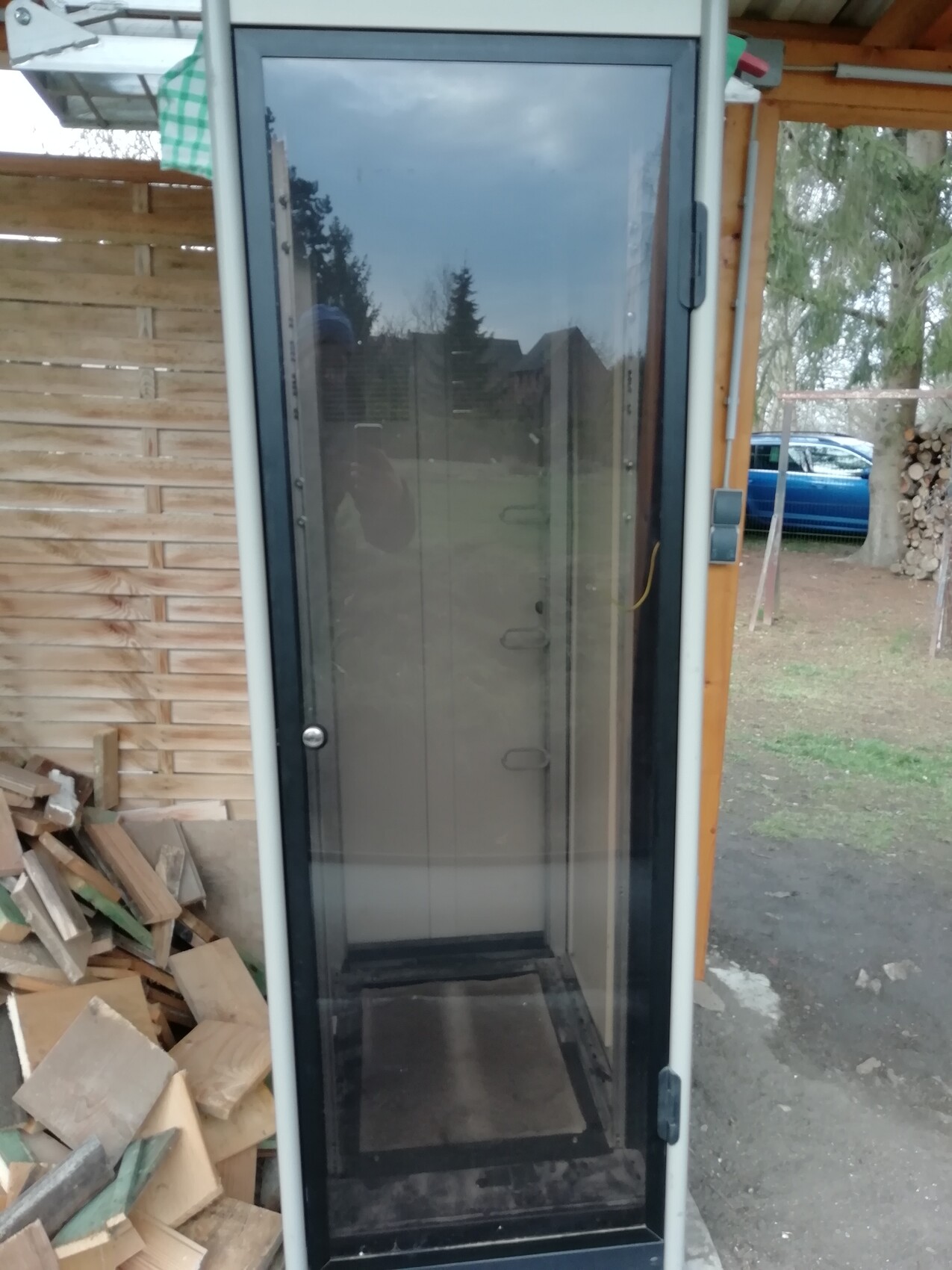 Freestanding glass display case outside next to stacks of wood; reflection of the photographer