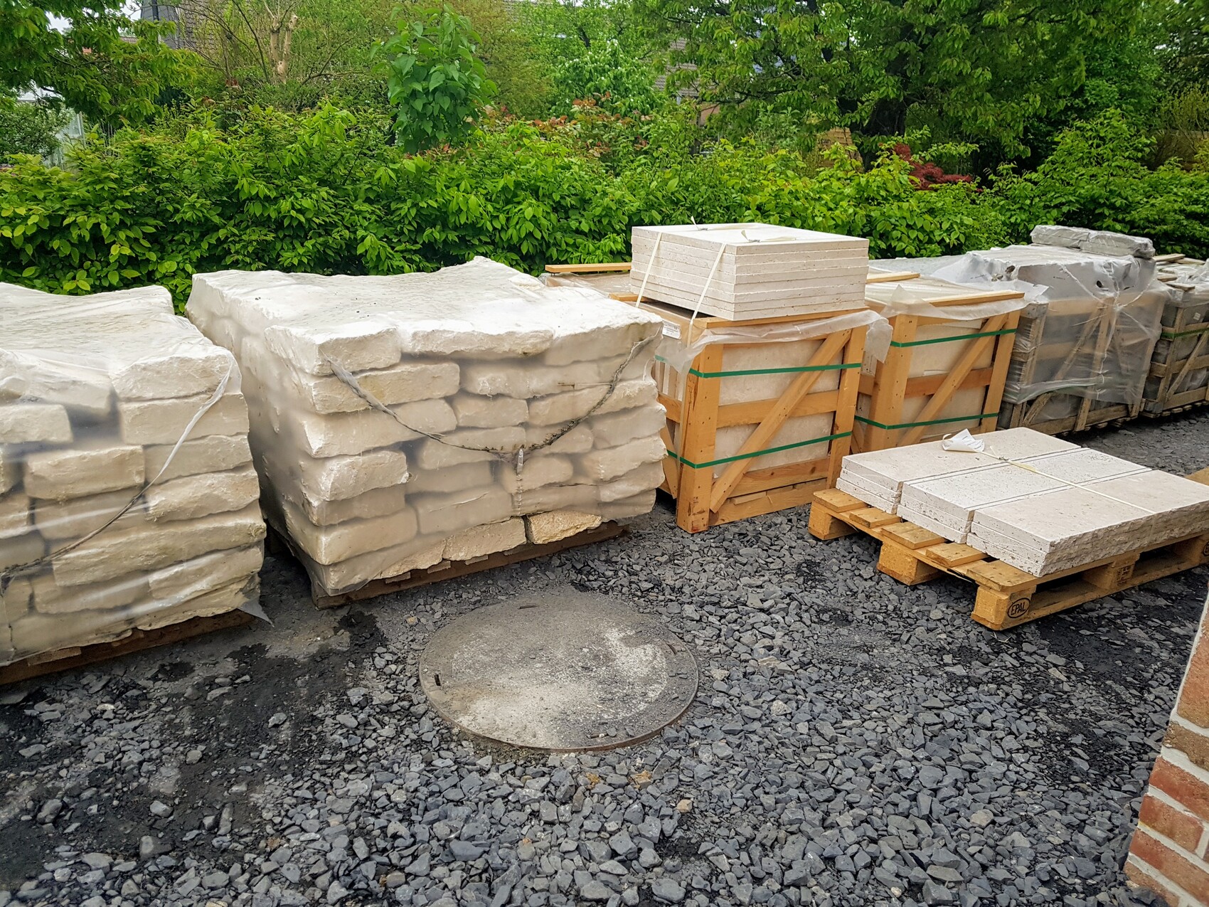 Large building material stacks of stone slabs and blocks on pallets in front of a green hedge.