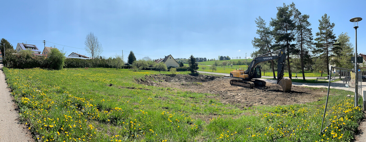 Excavator worksite in an open field next to a footpath; yellow flowers, houses in the background.