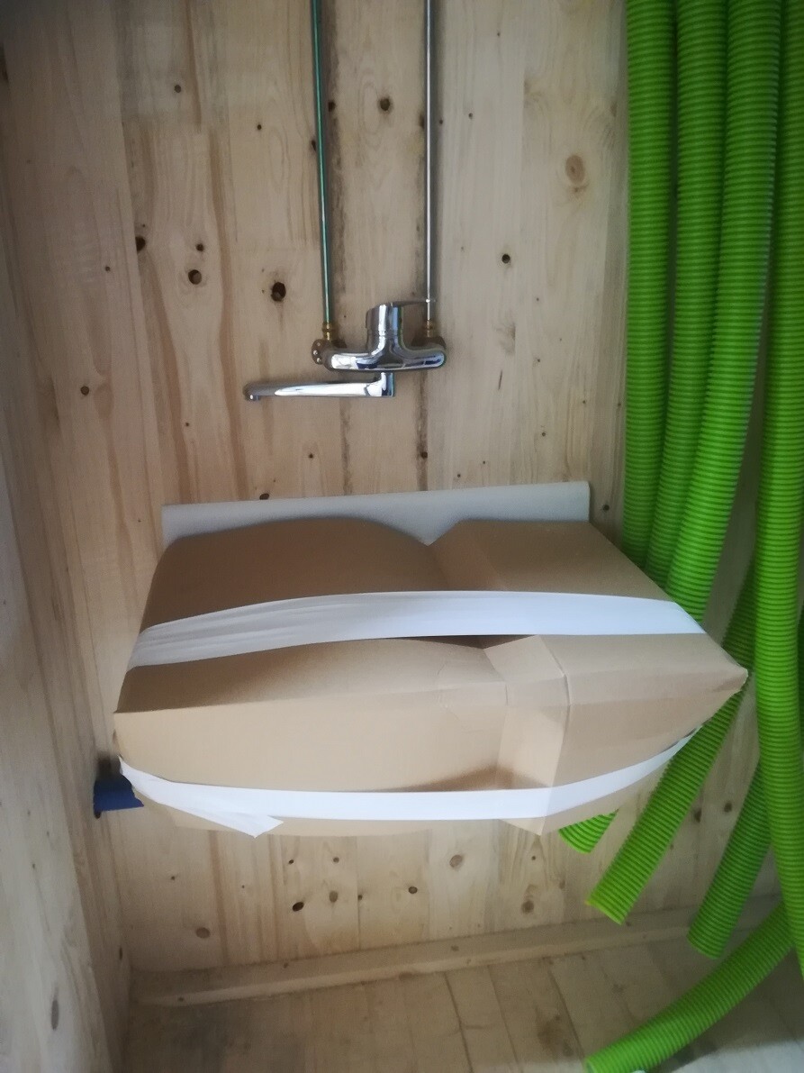 Chrome faucet on a wooden wall, brown cardboard secured with white tape, green hoses to the right.