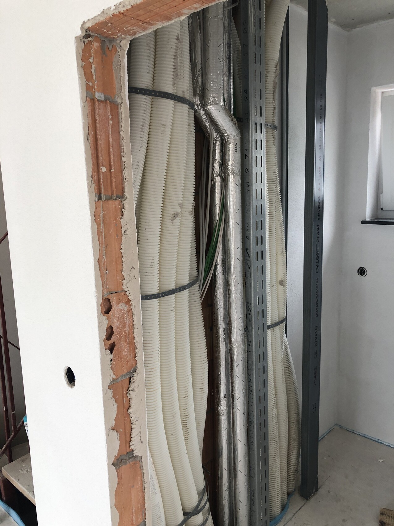 Unfinished wall with cable ducts, pipes and metal supports; cable bundles behind drywall.