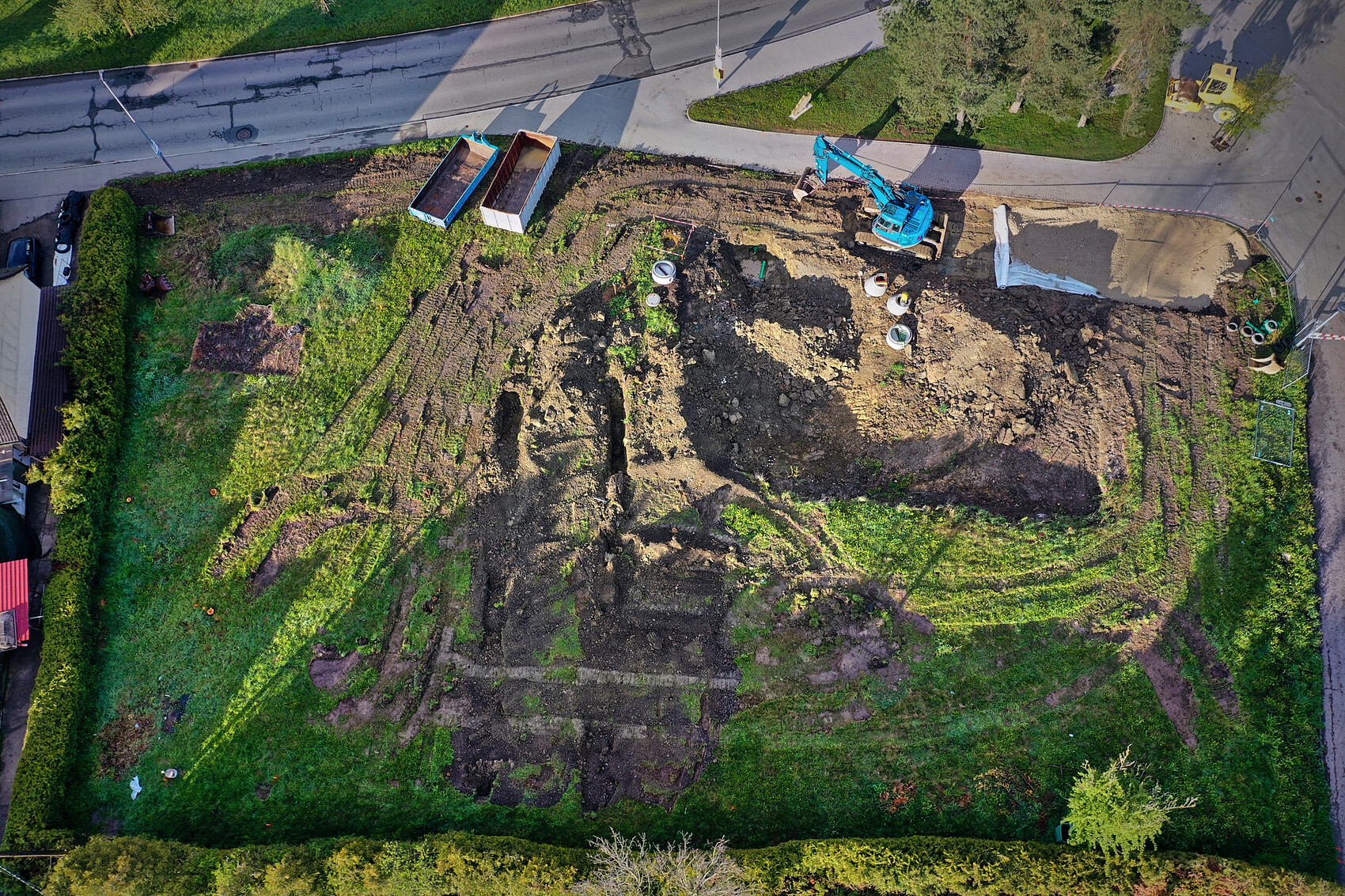 Aerial view of a construction site in the garden: blue excavator digging earth, container next to it.