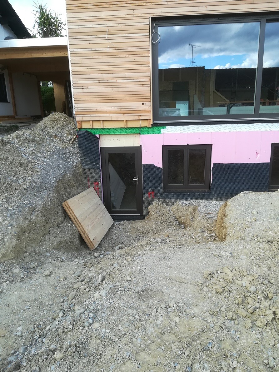 Construction site view: door and two windows in the basement, pink insulation boards, earthworks.